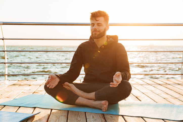 Relaxed young man meditating while sitting on a fitness mat at the beach